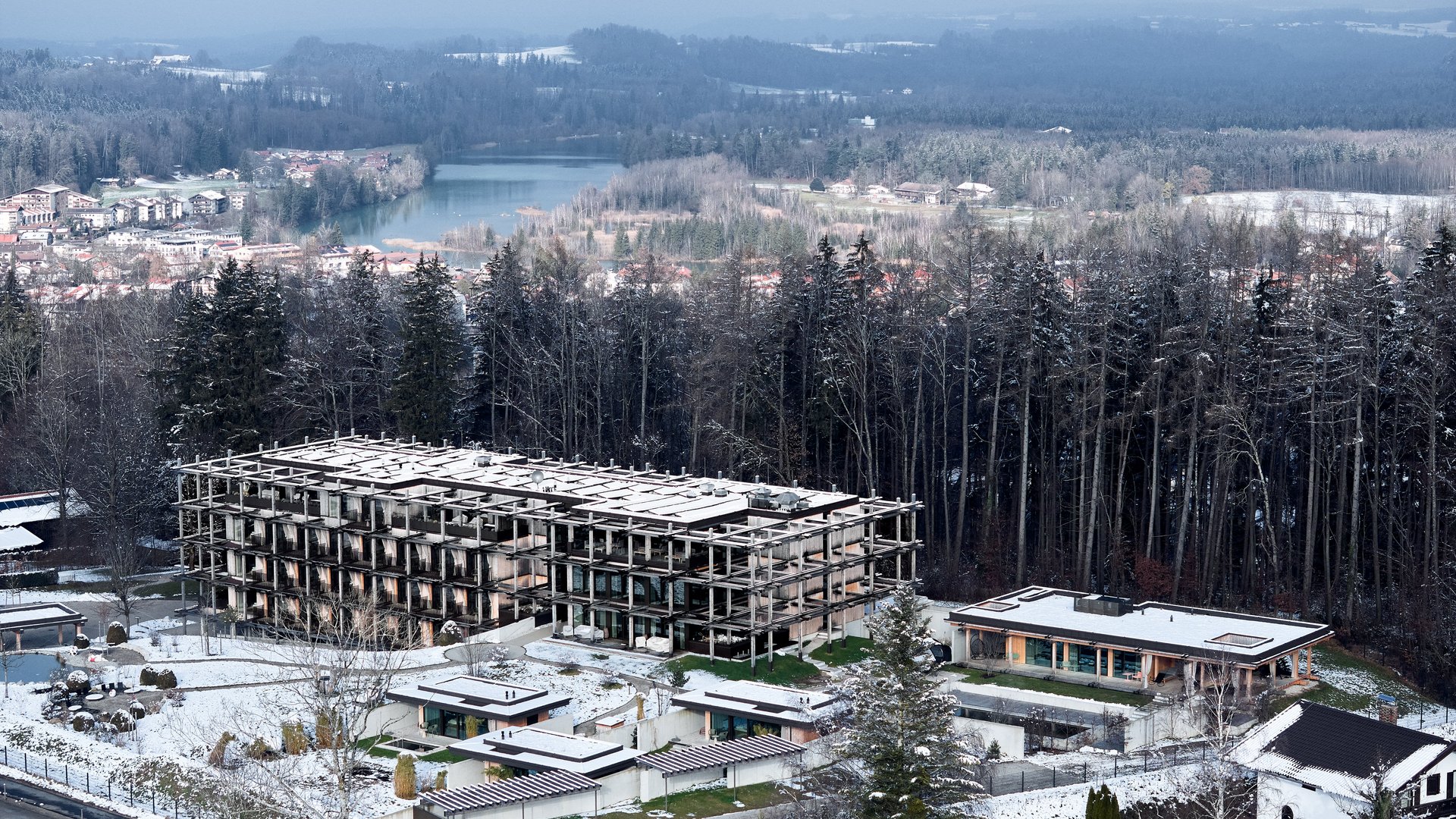 BERGEBLICK: Ihr Hotel in Bad Tölz Verschneite Gebäude am Waldrand, dahinter ein See und ein mehrstöckiger Rohbau