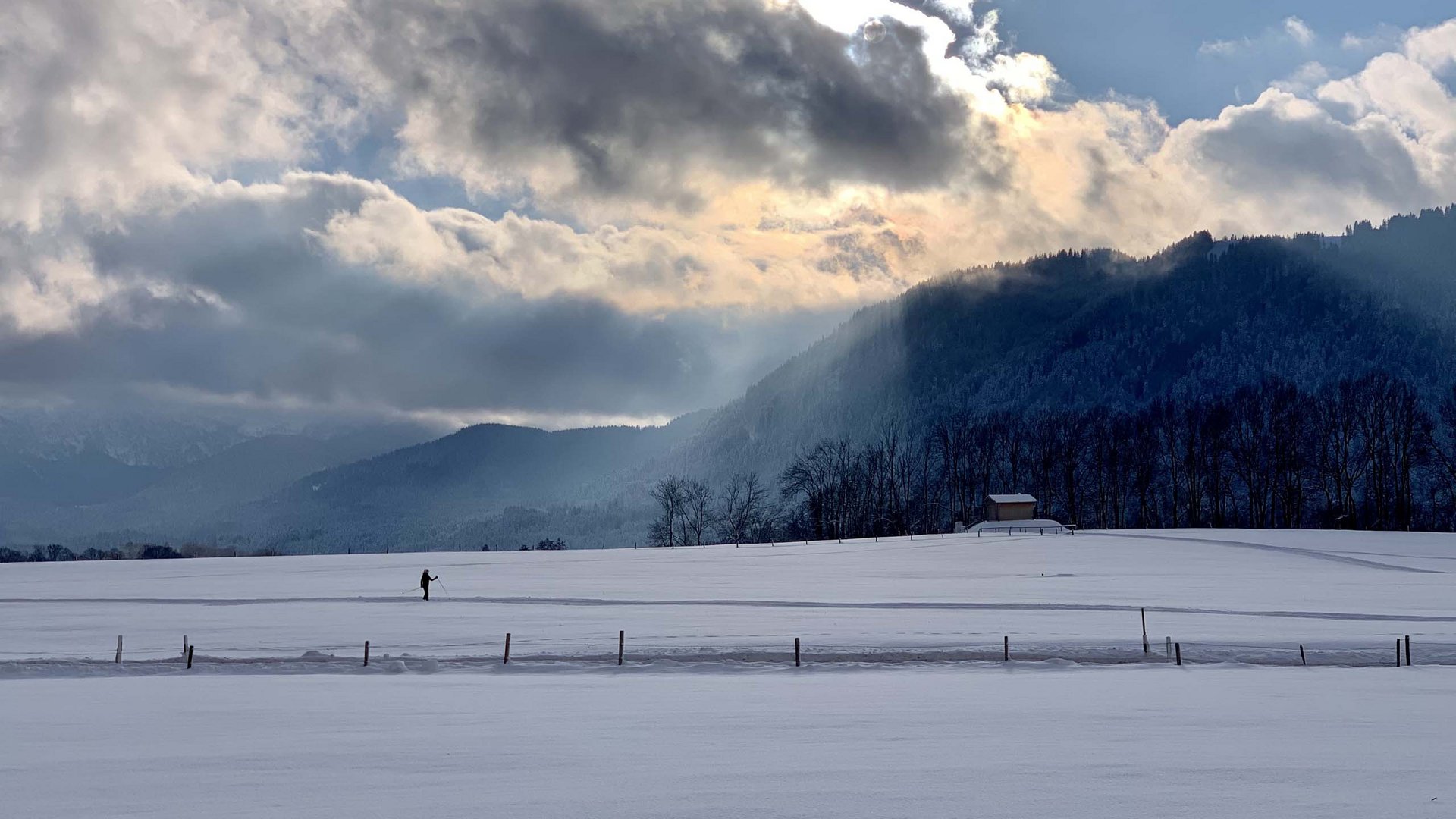 Dein Aktivurlaub in Bayern in Bad Tölz Landschaft mit verschneitem Feld, Bergen und einer Person beim Skilanglauf