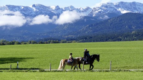 Aussicht auf Traumurlaub in Bad Tölz Menschen reiten Pferde auf einem Weg vor grünen Wiesen und schneebedeckten Bergen