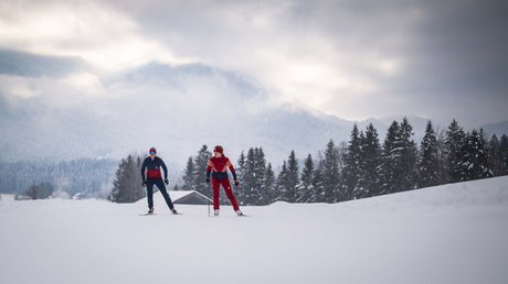 Aussicht auf Traumurlaub in Bad Tölz Zwei Skifahrer in roter und blauer Kleidung auf schneebedecktem Feld mit Bäumen und Bergen