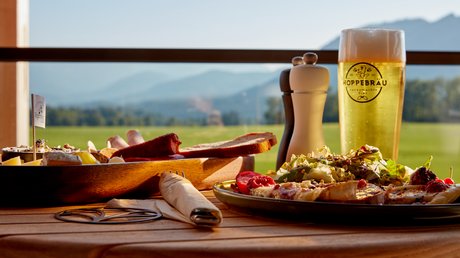 Aussicht auf Traumurlaub in Bad Tölz Bier, Brotzeitbrett und Salat auf einem Holztisch mit Bergblick