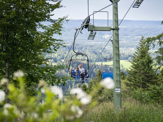 Dein Aktivurlaub in Bayern in Bad Tölz Mutter und Kind auf Sessellift im grünen Wald mit Bergpanorama