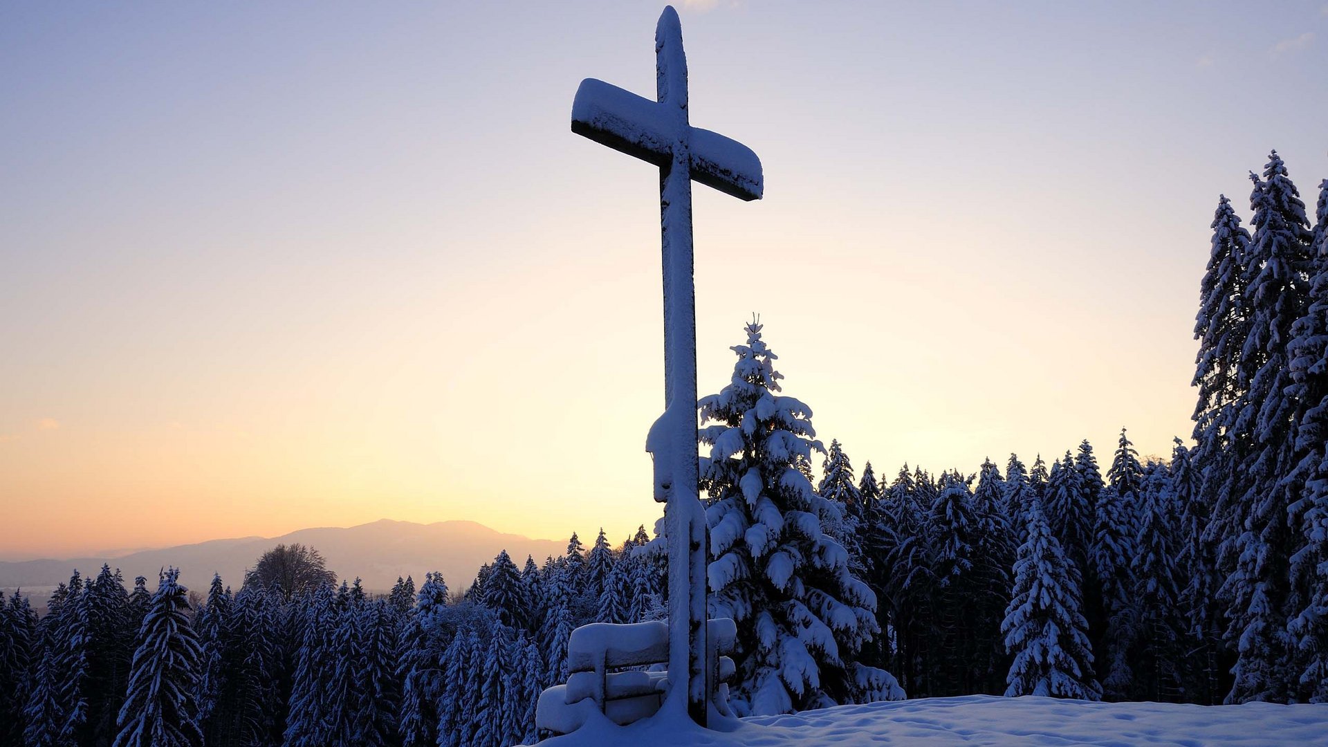 Dein Aktivurlaub in Bayern in Bad Tölz Schneebedecktes Kreuz auf einem Hügel mit verschneitem Wald bei Sonnenuntergang