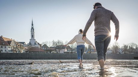Aussicht auf Traumurlaub in Bad Tölz Paar watet barfuß in einem Fluss vor einem Dorf mit Kirchturm bei Sonnenuntergang