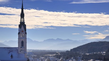 Aussicht auf Traumurlaub in Bad Tölz Kirchturm über schneebedecktem Dorf mit Bergen und blauem Himmel im Hintergrund