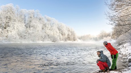 Aussicht auf Traumurlaub in Bad Tölz Zwei Personen in bunter Winterkleidung am verschneiten Flussufer unter blauem Himmel