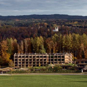 BERGEBLICK: Ihr Hotel in Bad Tölz Modernes Hotelgebäude vor herbstlichem Wald und Hügeln mit Schloss im Hintergrund